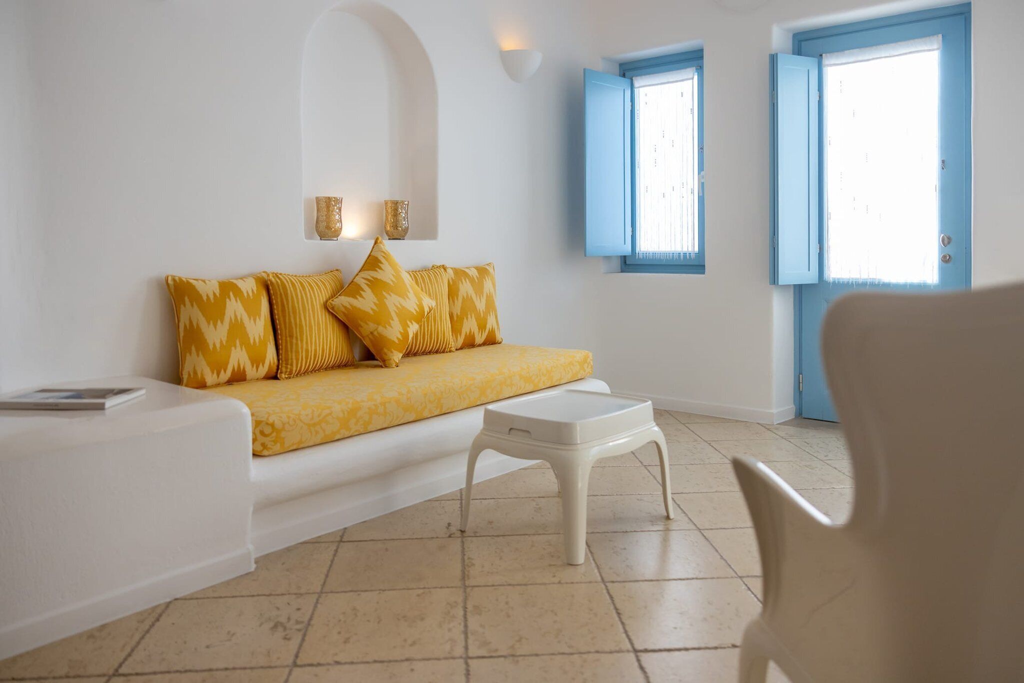 A room with a sofa, a small table, and a chair. There are two windows with blue shutters and a door with a blue frame.