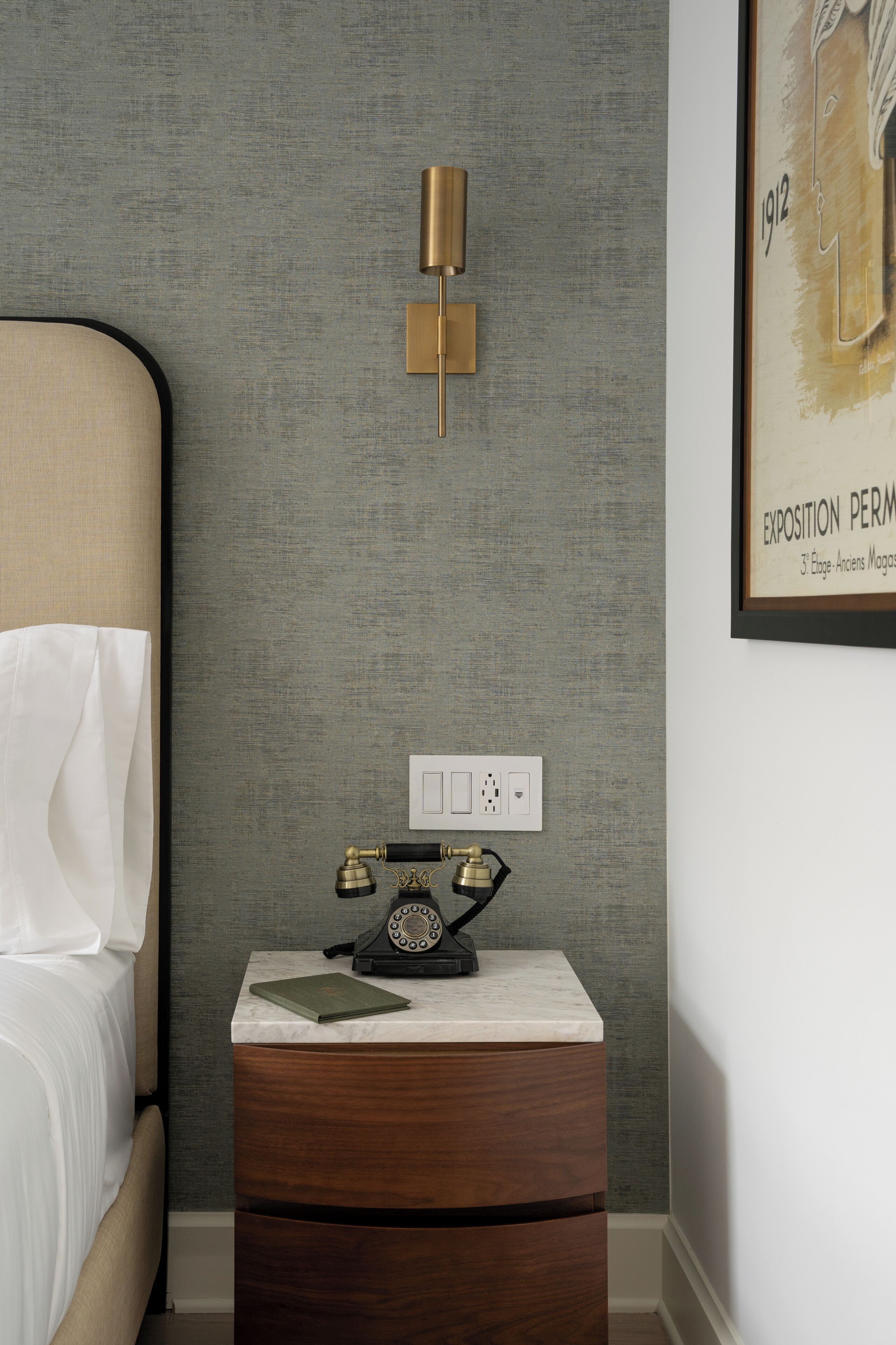 A bedroom with a bed, a nightstand with a telephone, a wall-mounted light fixture, and a framed picture on the wall.