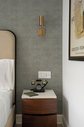 A bedroom with a bed, a nightstand with a telephone, a wall-mounted light fixture, and a framed picture on the wall. - Manoir Émery (Montréal)