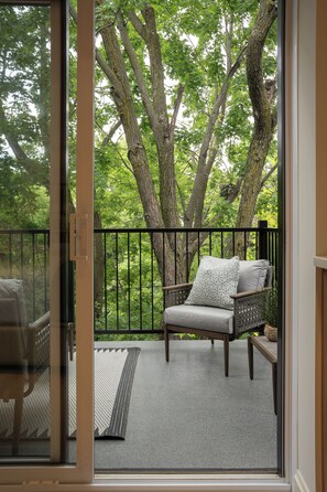 A balcony with a wicker chair and a cushion, a small rug, and a view of trees through a glass door. - Manoir Émery (Montréal)