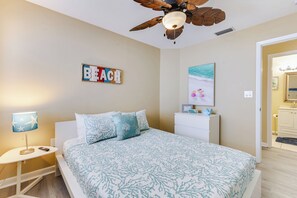 Ein Schlafzimmer mit Bett, Deckenventilator, Nachttisch, Lampe, einer Wandtafel, einem Gemälde und einer Badezimmertür.