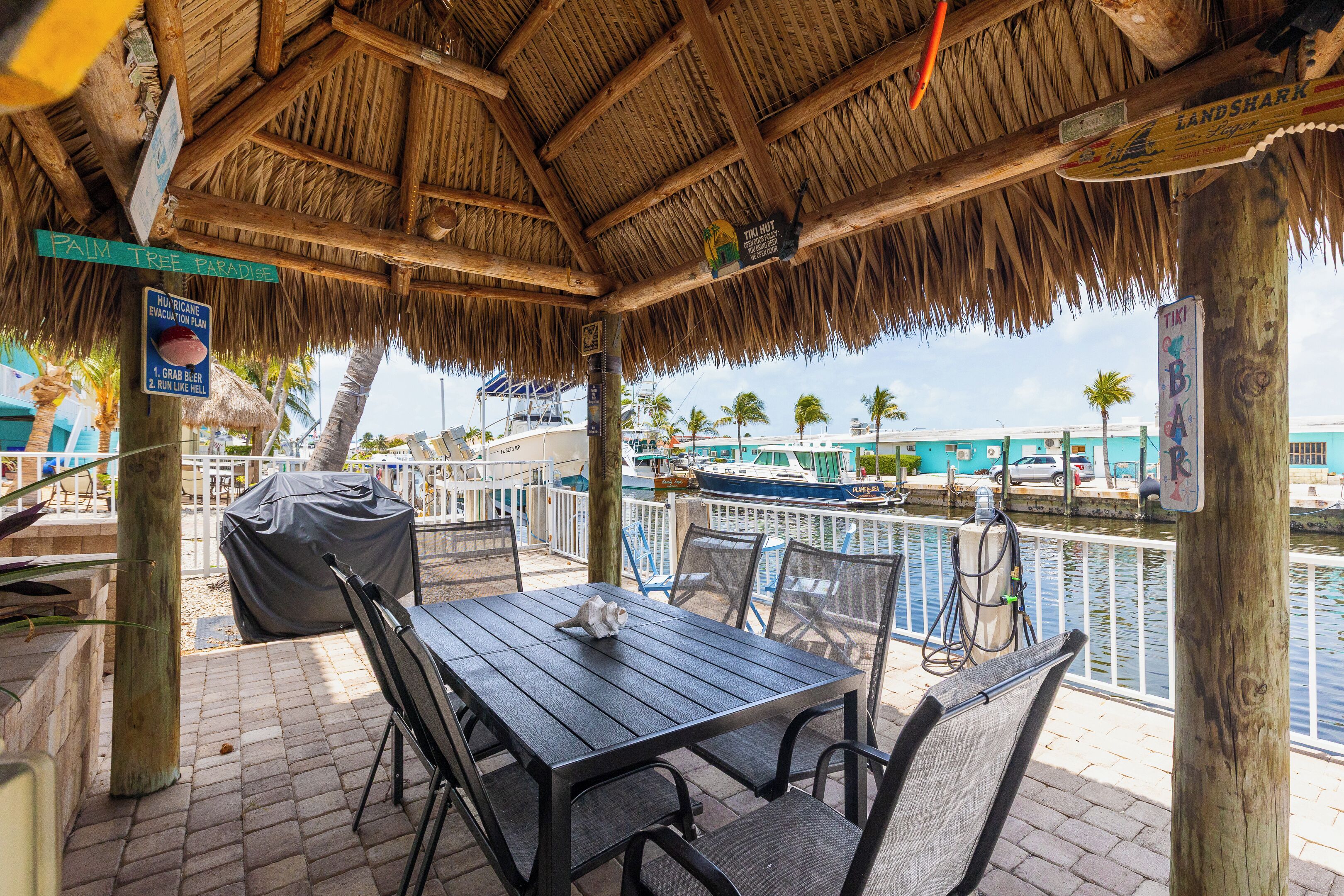 An outdoor seating area with a thatched roof, a table, and chairs, overlooking a water body with boats.