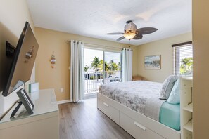 A bedroom with a bed, a ceiling fan, a TV on a glass table, and a view of palm trees through the window.
