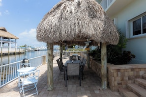 A covered outdoor seating area with a thatched roof, a table, and chairs, overlooking a marina with boats.