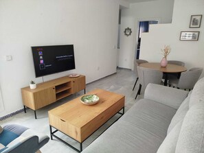 A modern living room with a flat-screen TV, wooden coffee table, grey sofa, and a dining area with a round table and chairs. - Apartment near a magical spot in Chiclana (Poblado de Sancti Petri)