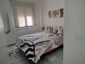 Une chambre à coucher avec un lit, une fenêtre avec des stores, une œuvre d’art accrochée au mur et une petite table de chevet.
