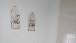 En säng med en vit madrass och en blå handduk, ett litet runt bord med en vit lampa och två väggdekorationer med surftema.