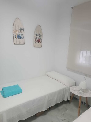 Ein Einzelbett mit weißer Matratze und blauem Handtuch, ein kleiner runder Tisch mit einer weißen Lampe sowie zwei Wanddekorationen zum Thema Surfen.