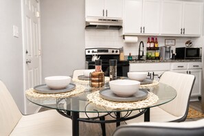 Ett modernt kök med vita skåp, ett matbord i glas med underlägg och en avdelning för kaffebryggning.