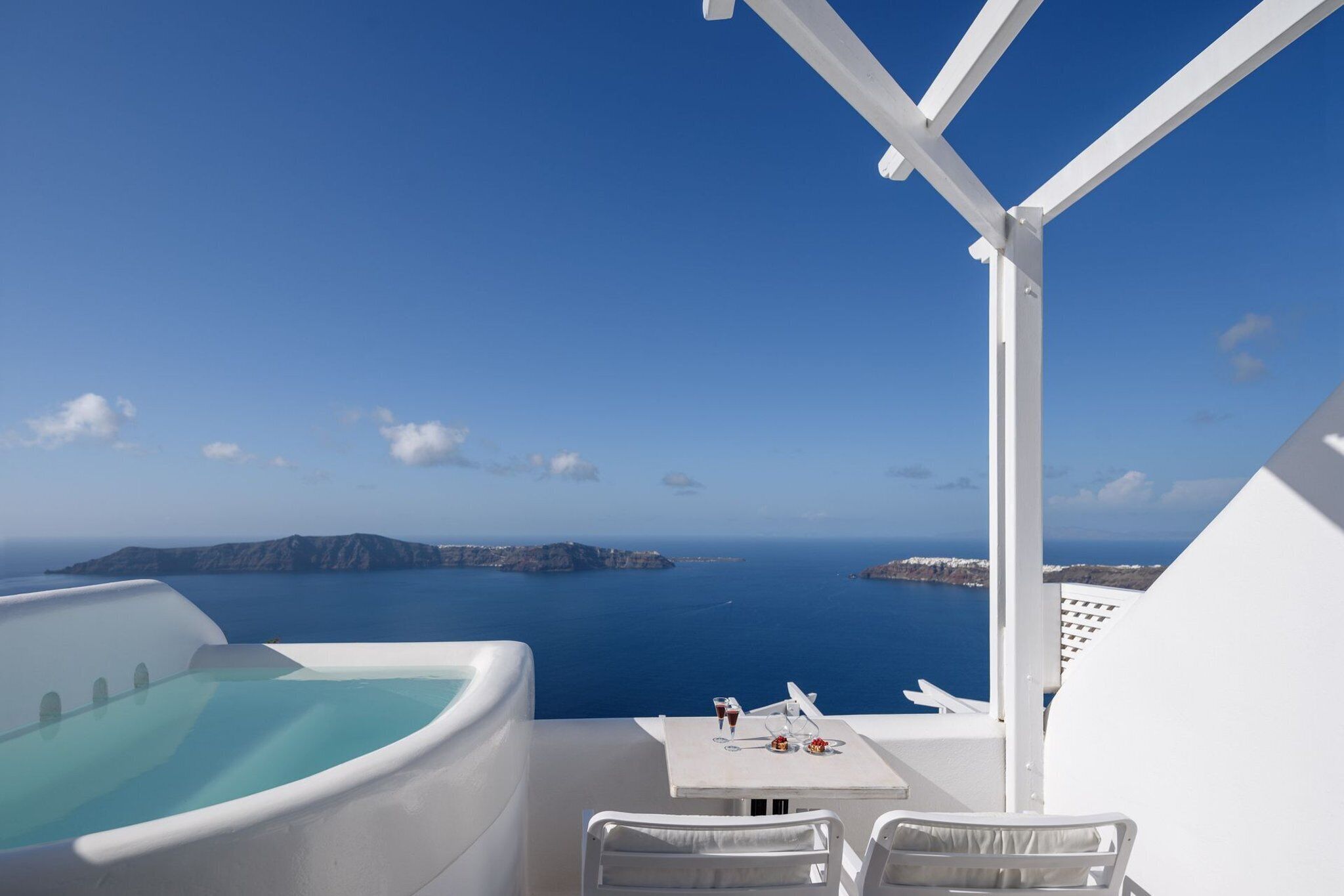 Un balcon avec vue sur la mer, une baignoire blanche et une table avec un verre de vin et une assiette de nourriture.