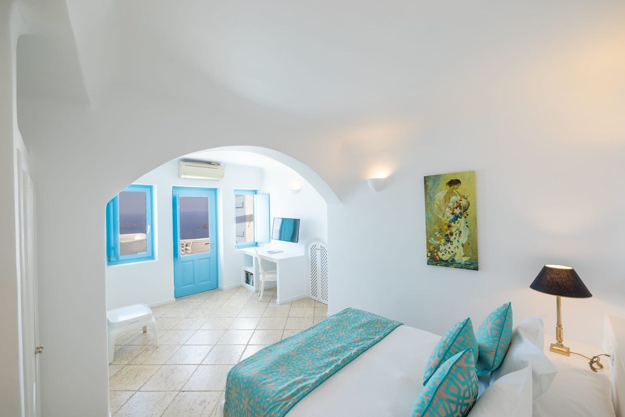 Une pièce comprenant un lit, un bureau et une chaise, une porte en arc donnant sur une autre pièce, et un tableau accroché au mur.