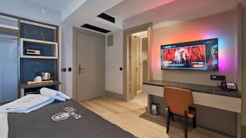 A modern hotel room with a bed, a desk, a chair, a TV, and a minibar.