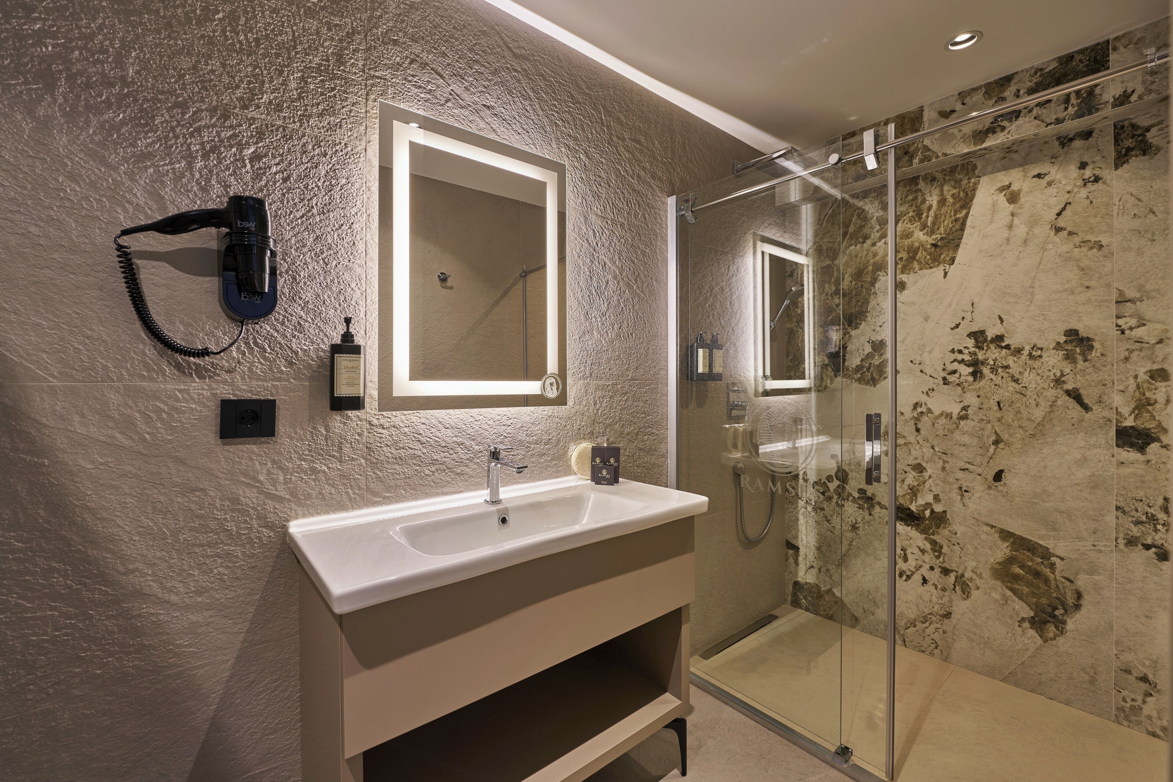 Ein modernes Badezimmer mit einem weißen Waschbecken, einem an der Wand montierten Haartrockner, einer Duschkabine aus Glas und einer strukturierten Wand.