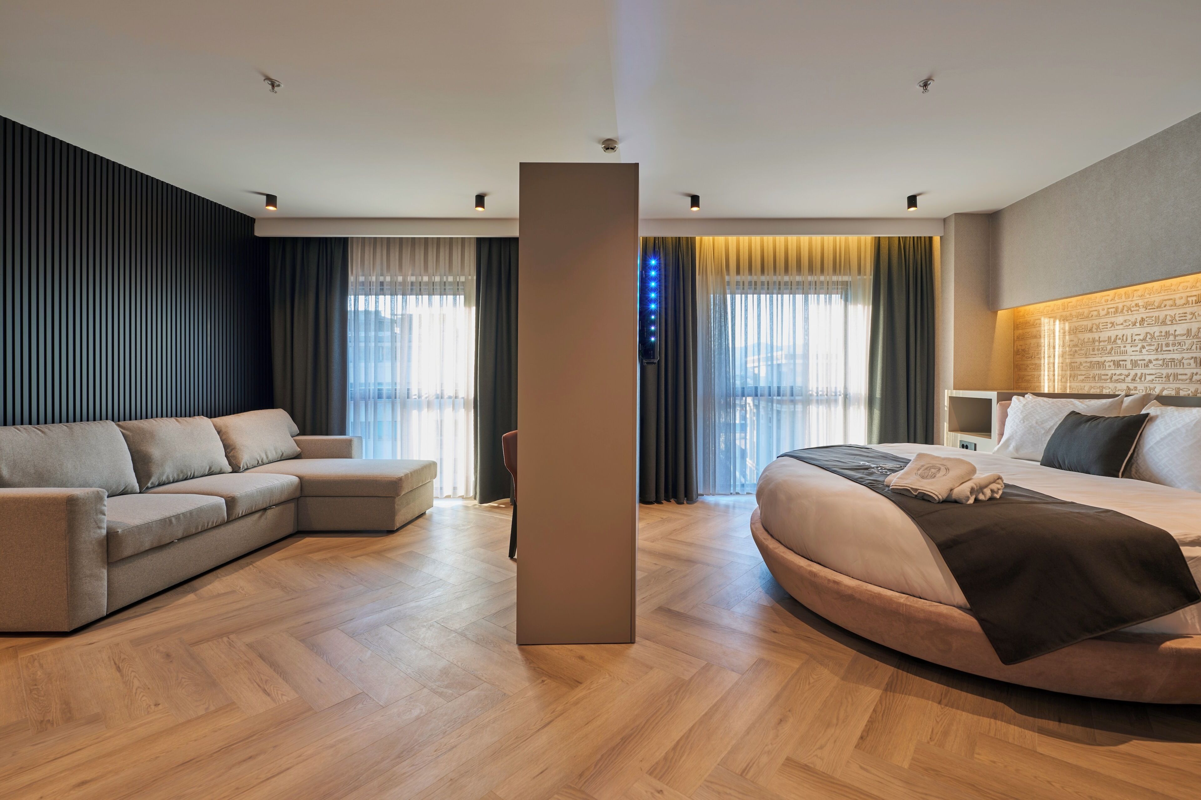 A modern hotel room with a sofa, a bed, a wooden floor, and a view of the city through the windows.