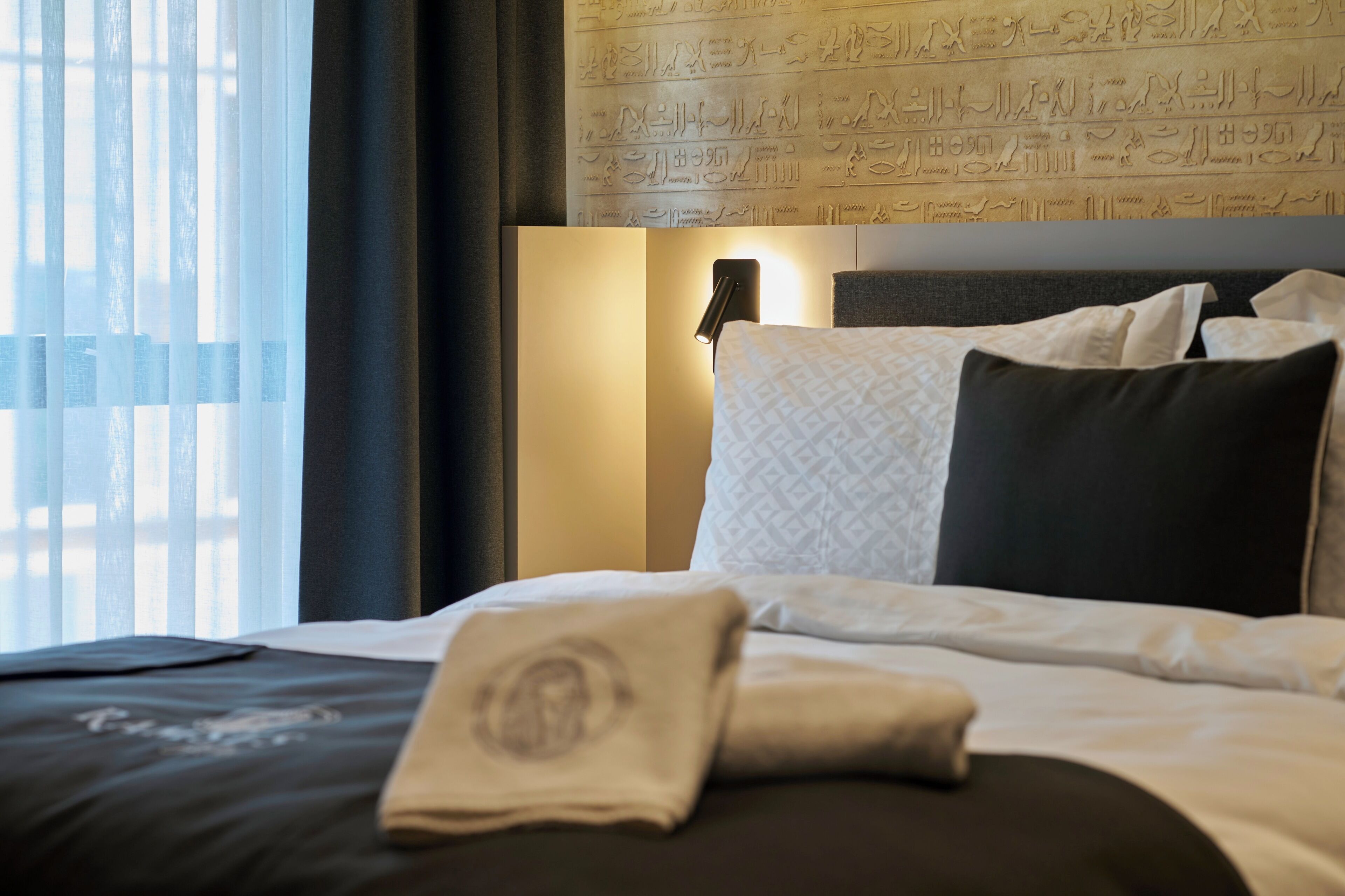 A hotel room with a bed, pillows, a nightstand, a lamp, and a wall with hieroglyphs.