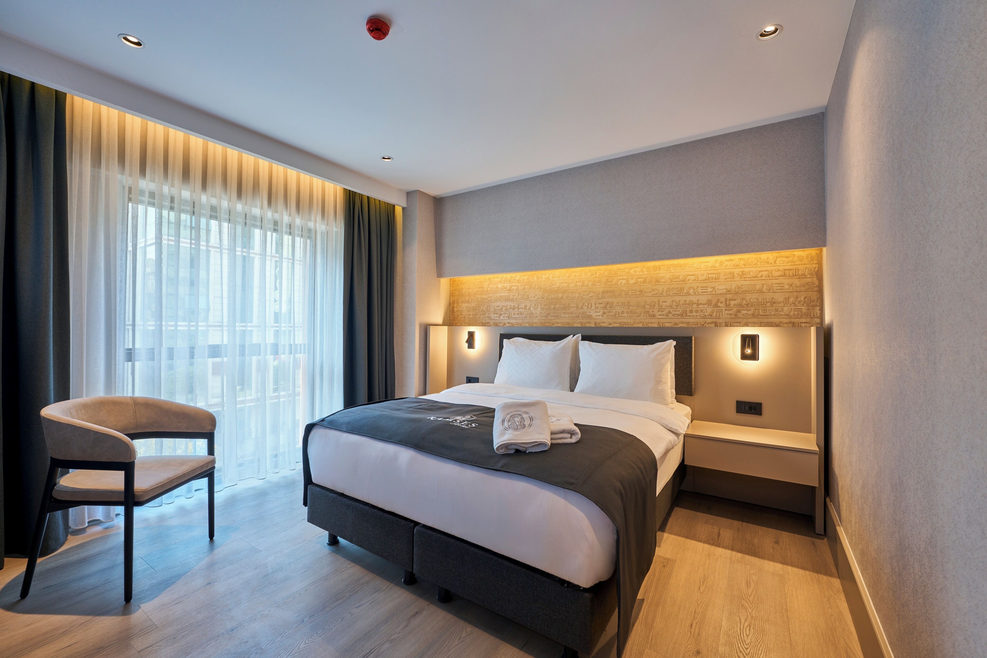 Une chambre d’hôtel comprenant un lit, une chaise, une table de chevet et un luminaire fixé au mur.