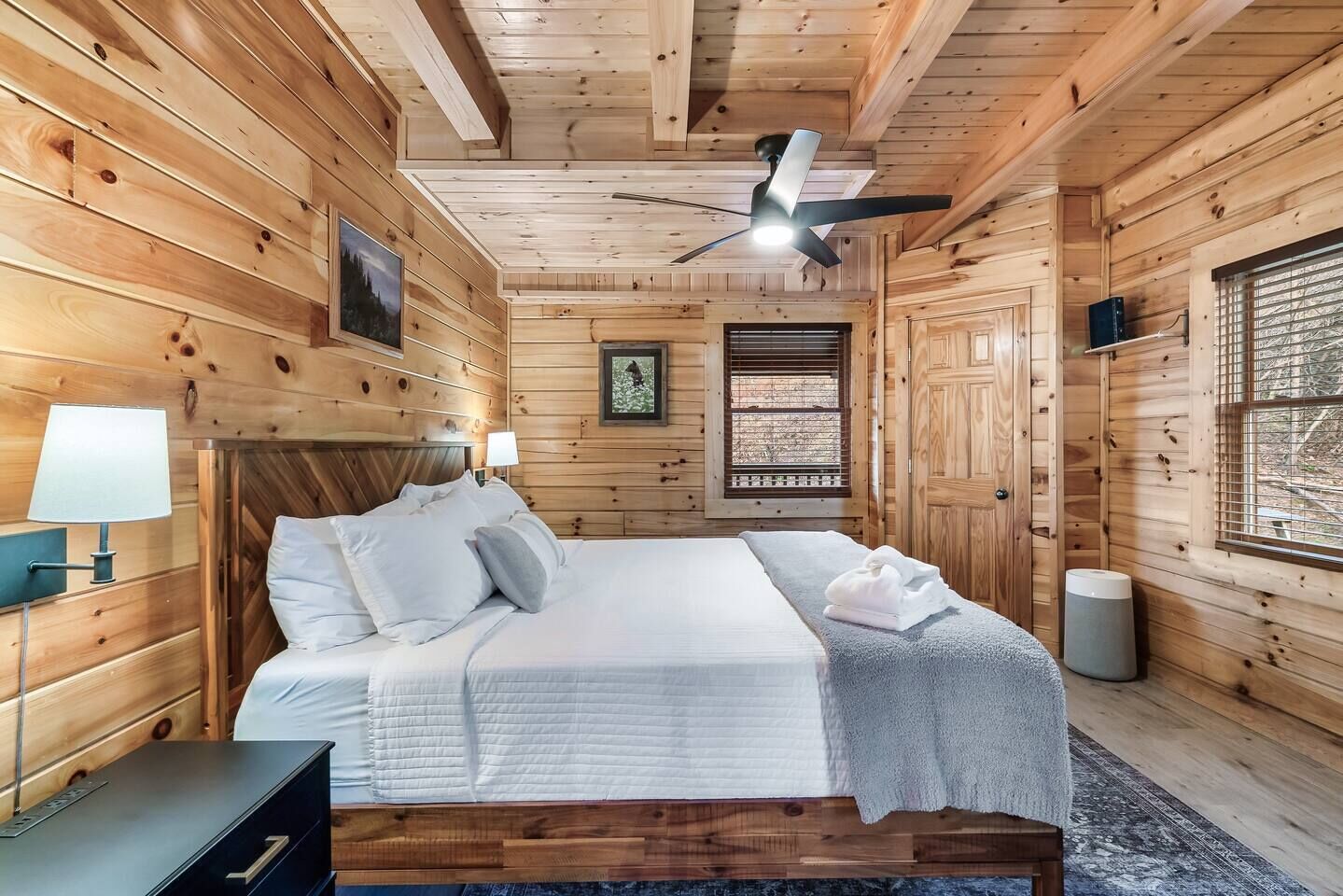 Une chambre à coucher dans une cabane en bois, avec un lit, des lampes de chevet, un ventilateur de plafond et une fenêtre avec des stores.