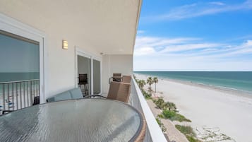 Un balcon avec une table et des chaises en verre, donnant sur une plage bordée de palmiers et offrant une vue sur un ciel dégagé.
