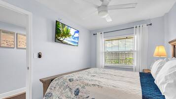 Une chambre à coucher avec un lit, un ventilateur de plafond, une fenêtre avec des stores et un téléviseur fixé au mur.