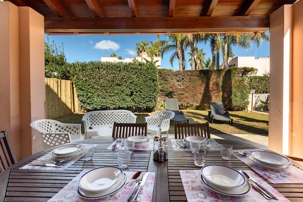Un espace de restauration en plein air avec une table en bois prévue pour quatre personnes, entourée de chaises blanches, sous une pergola en bois.