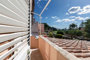 Un balcon d’habitation doté d’un toit en tuiles, d’une balustrade blanche et offrant une vue dégagée sur la rue et la verdure en contrebas.