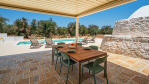 Un espace extérieur abrité, aménagé pour les repas, avec une piscine, des murs en pierre et des oliviers en arrière-plan.