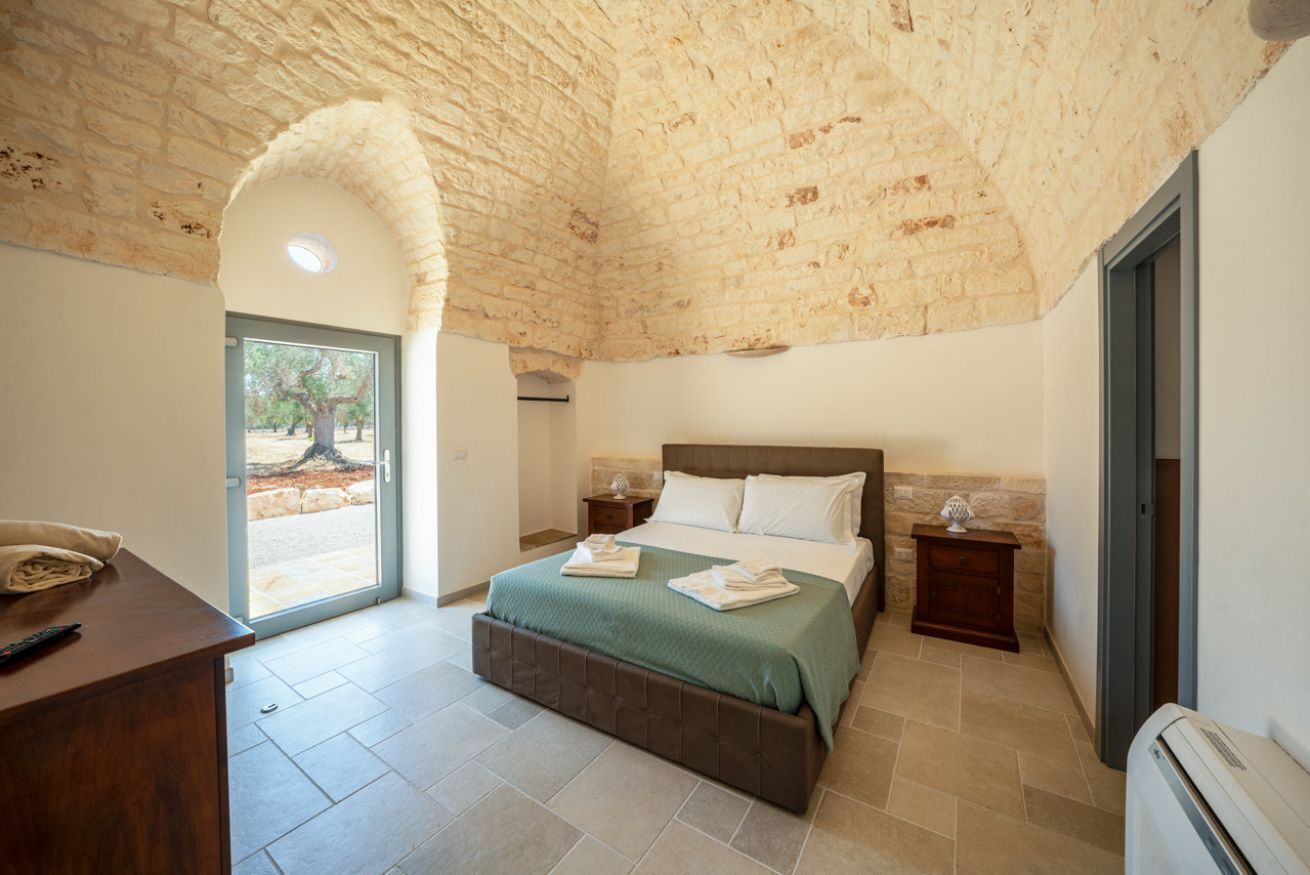 A bedroom with a stone ceiling, a bed with a green bedspread, a desk with a remote control, and a door leading outside.