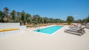 Une piscine à l’eau d’un bleu limpide, entourée de chaises longues et bordée d’un mur blanc agrémenté d’un coussin jaune.
