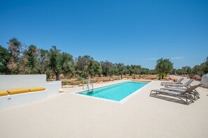 Ein Pool mit klarem, blauem Wasser, umgeben von Liegestühlen und einer weißen Wand mit einem gelben Kissen.