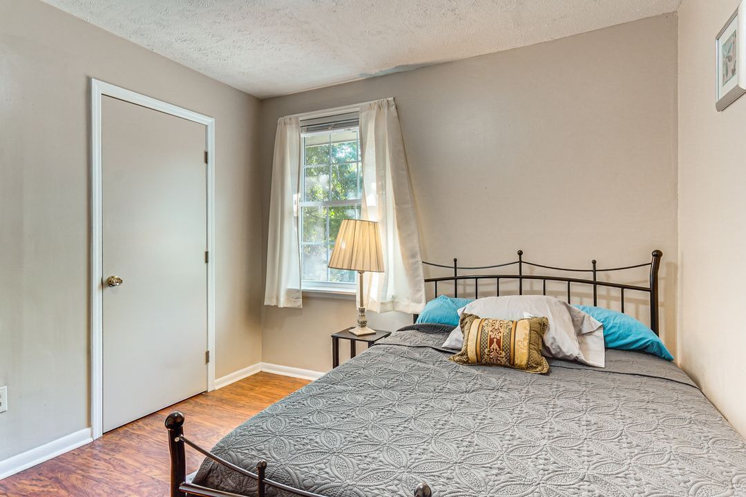 Un dormitorio con cama, ventana con cortinas, mesita de noche con lámpara y puerta.