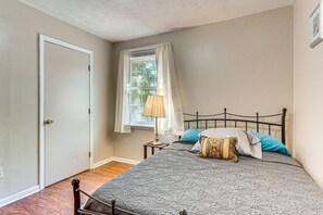 Ein Schlafzimmer mit einem Bett, einem Fenster mit Vorhängen, einem Nachttisch mit Lampe und einer Tür.