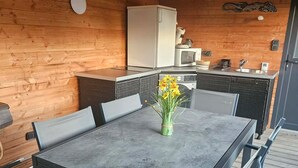 Un intérieur en bois comprenant une table à manger, des chaises, un coin cuisine et un plafond avec des poutres apparentes et des lampes suspendues.