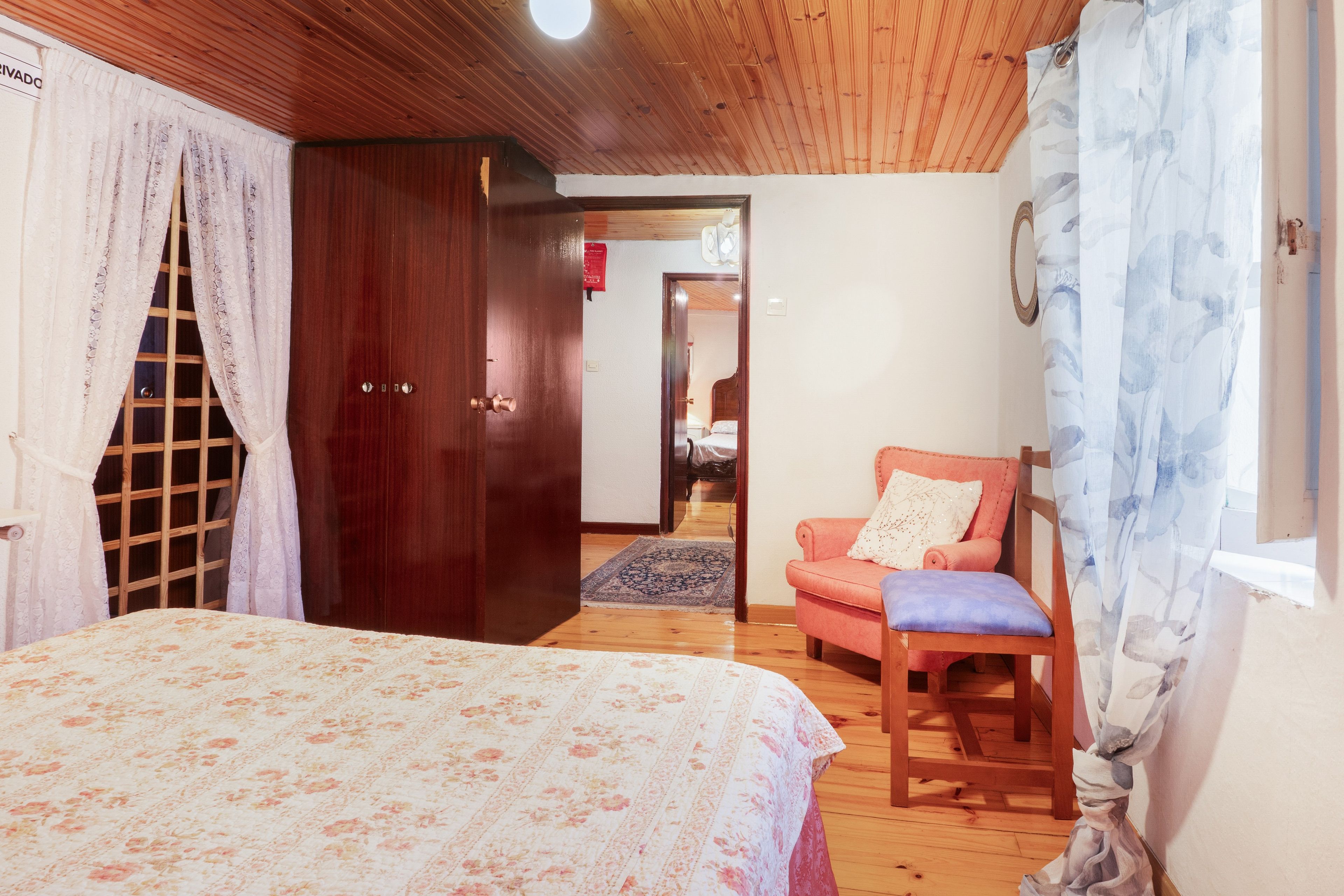 Un dormitorio con cama, armario de madera, una silla y una ventana con cortinas.