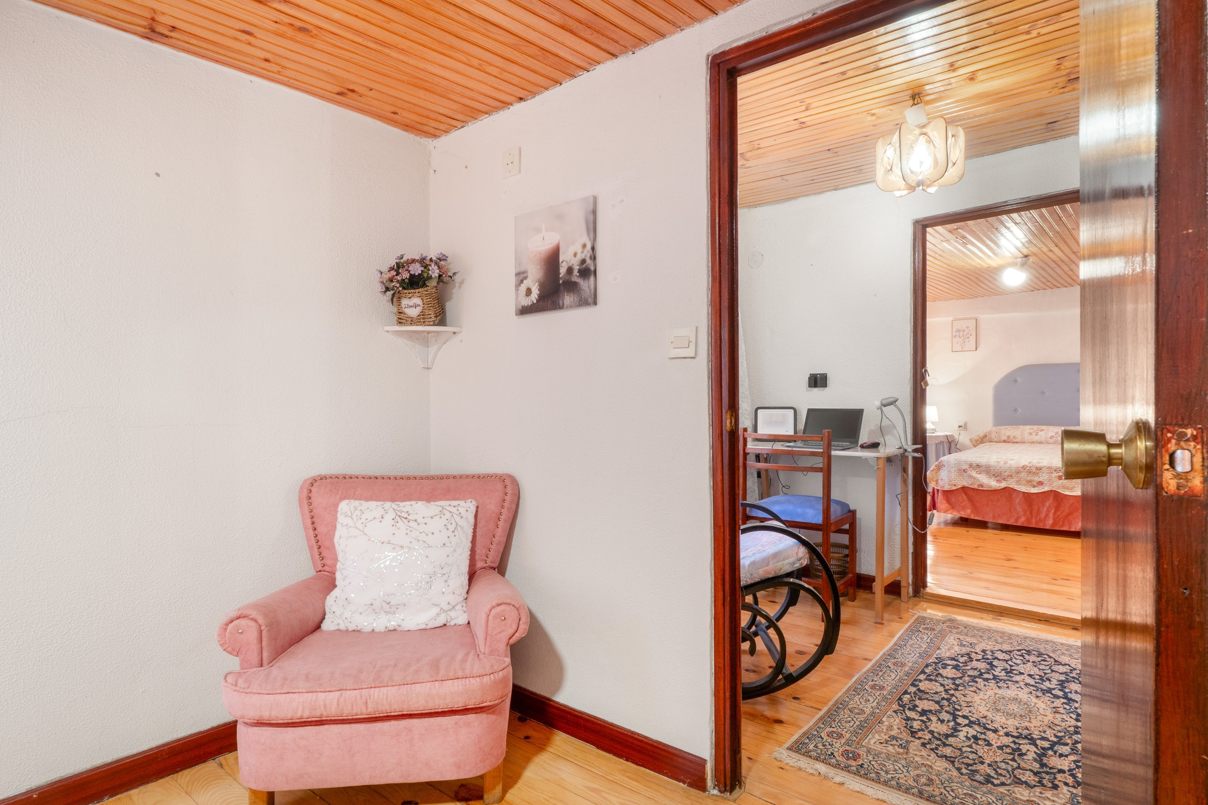 Ein Zimmer mit einem rosafarbenen Sessel, einem kleinen Regal mit einem Korb, einem Schreibtisch mit einem Laptop und einem Bett mit einem gemusterten Überwurf.