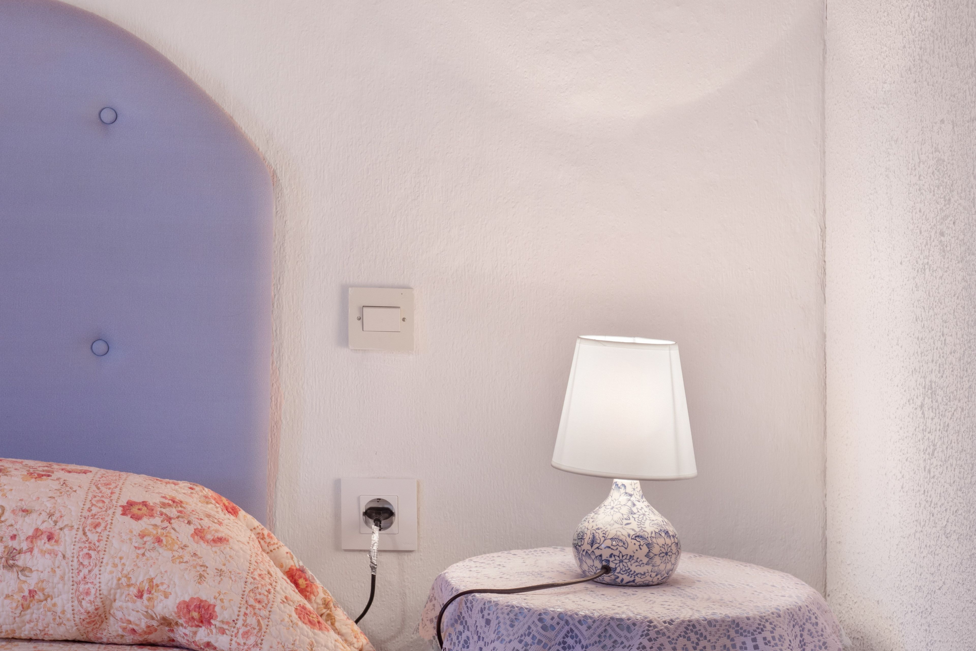 Un dormitorio con una cama, una mesita de noche con lámpara, un enchufe y un tomacorriente.