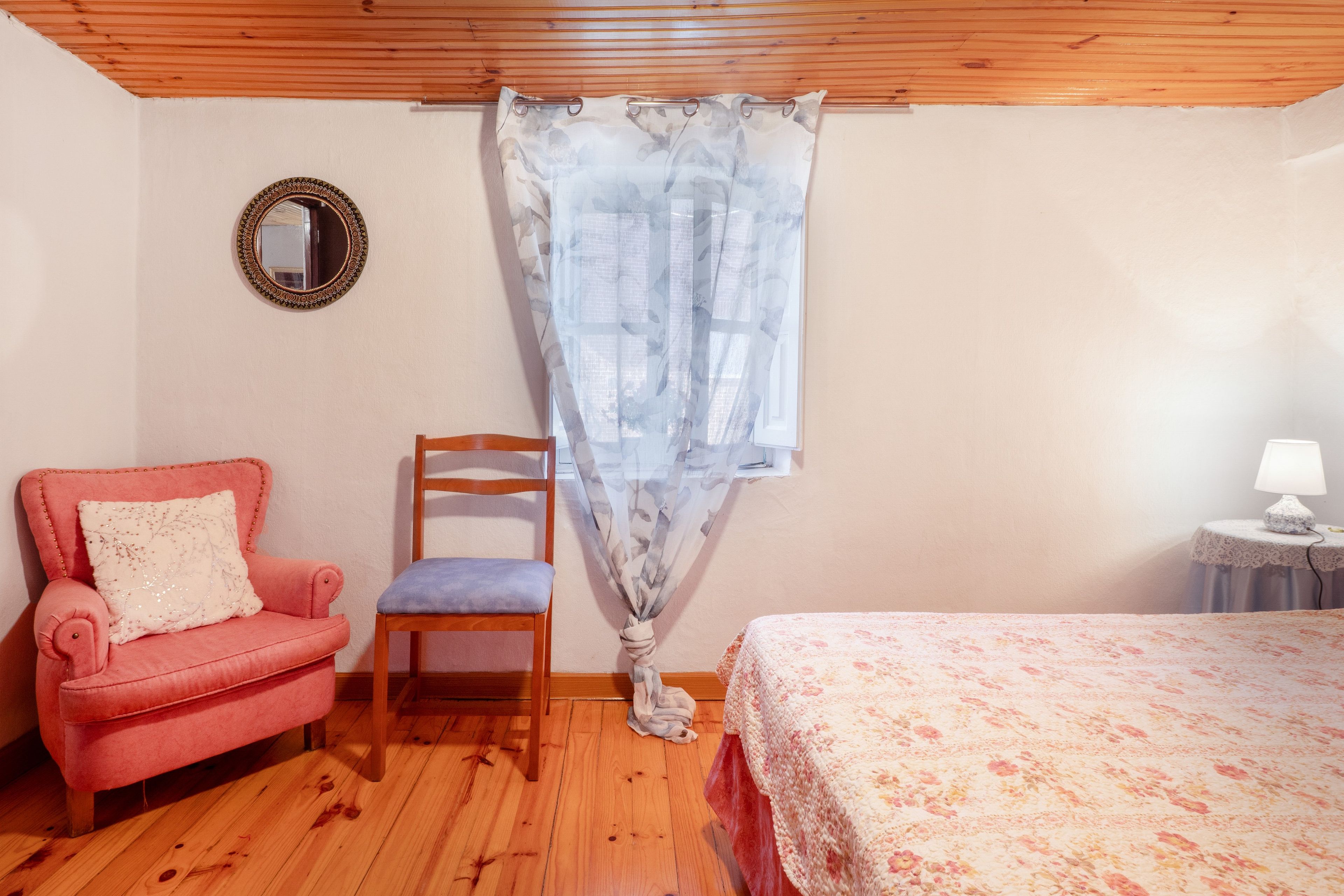Un dormitorio con techo de madera, colcha estampada de flores, sillón rosa, silla de madera y ventana con cortina translúcida.