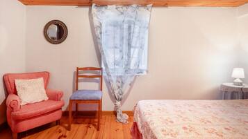 Un dormitorio con techo de madera, colcha estampada de flores, sillón rosa, silla de madera y ventana con cortina translúcida.