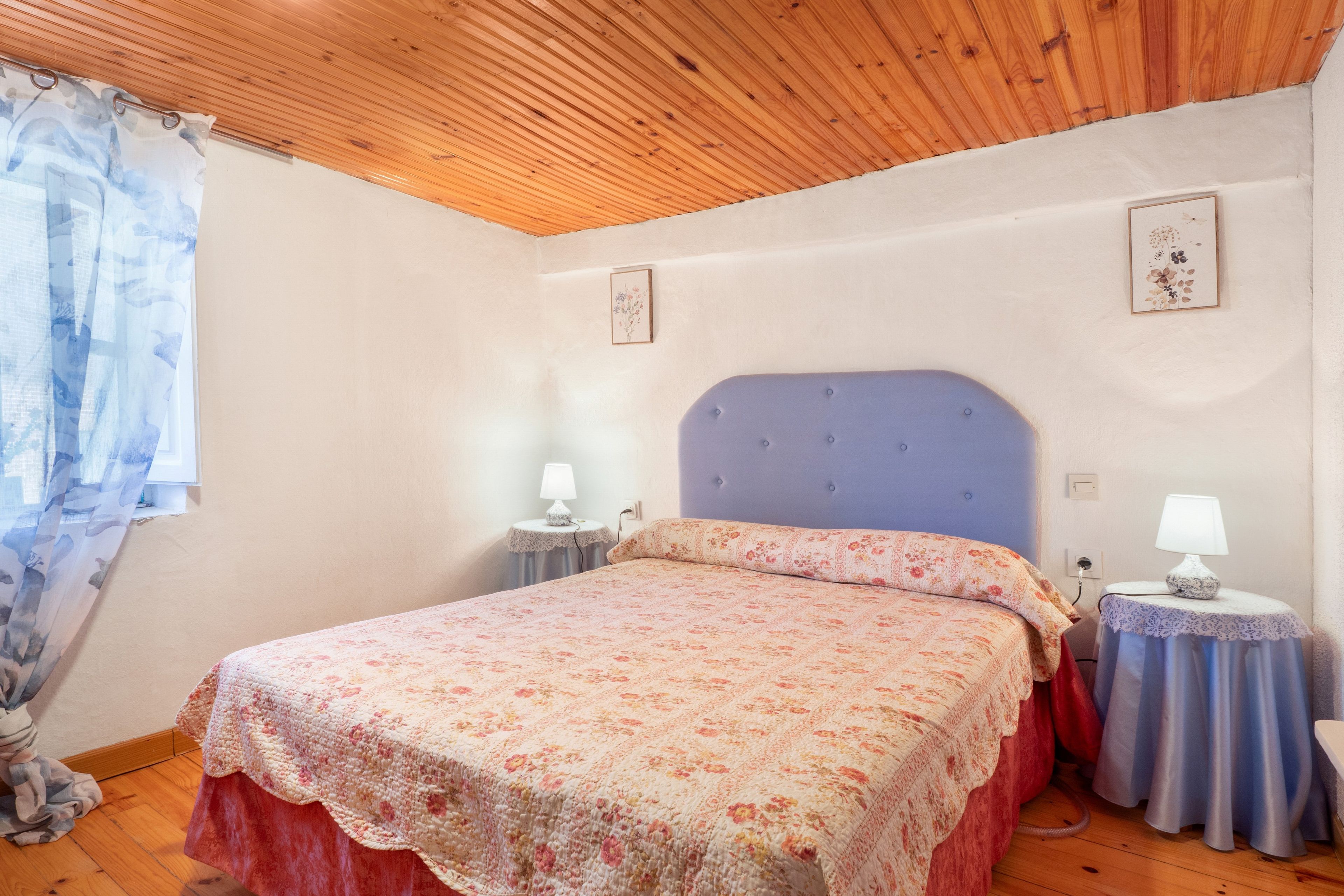 Un dormitorio con una cama, dos mesitas de noche con lámparas, un ventanal con cortinas y un techo de madera.
