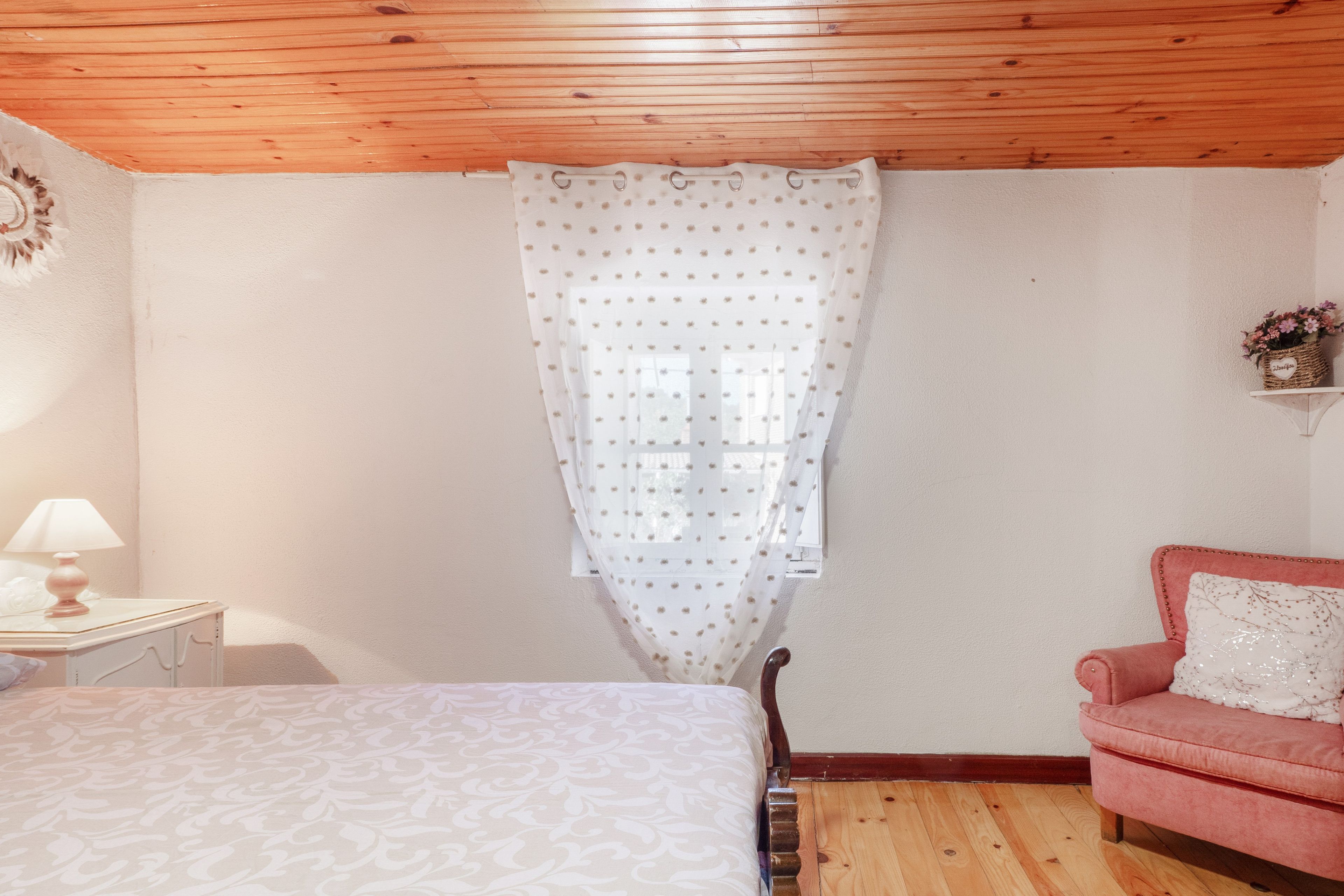 Un dormitorio con una cama, una silla, una mesita de noche, un ventanal con cortina y techo de madera.