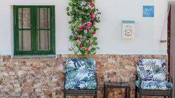 Een stenen muur met een groene deur, een wandbevestigde brievenbus en twee rieten stoelen met bloemkussens.