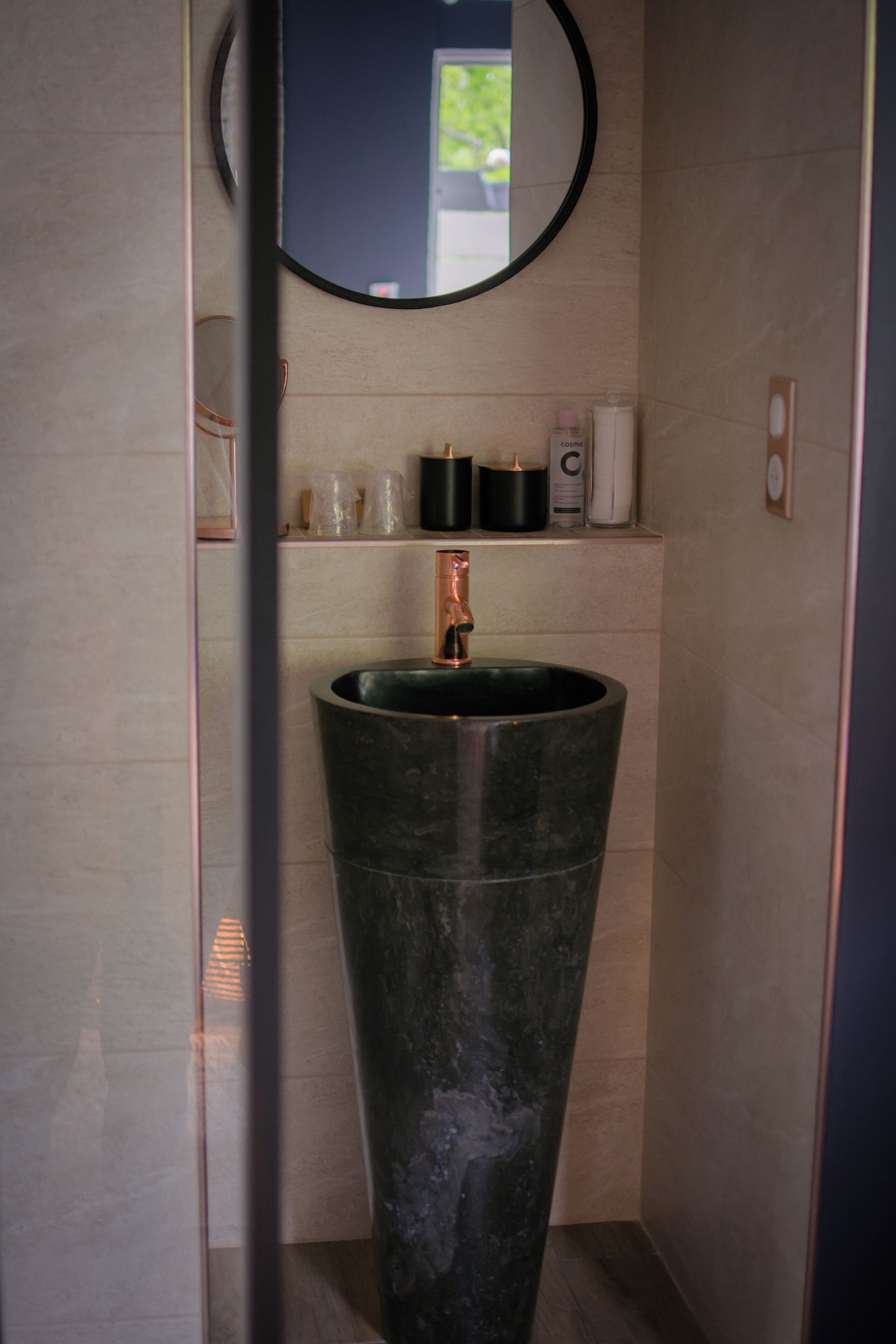 A bathroom with a large, dark-colored sink, a round mirror above it, and a shelf with toiletries.