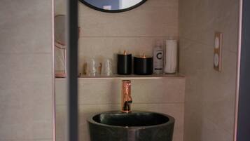 A bathroom with a large, dark-colored sink, a round mirror above it, and a shelf with toiletries.