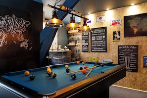 Une table de billard avec des billes, un éclairage suspendu et divers panneaux et affiches accrochés aux murs.