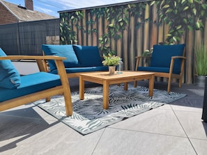 Une terrasse avec une table en bois et des chaises rembourrées bleues, un tapis à motifs et un mur orné d’une fresque représentant des feuillages.