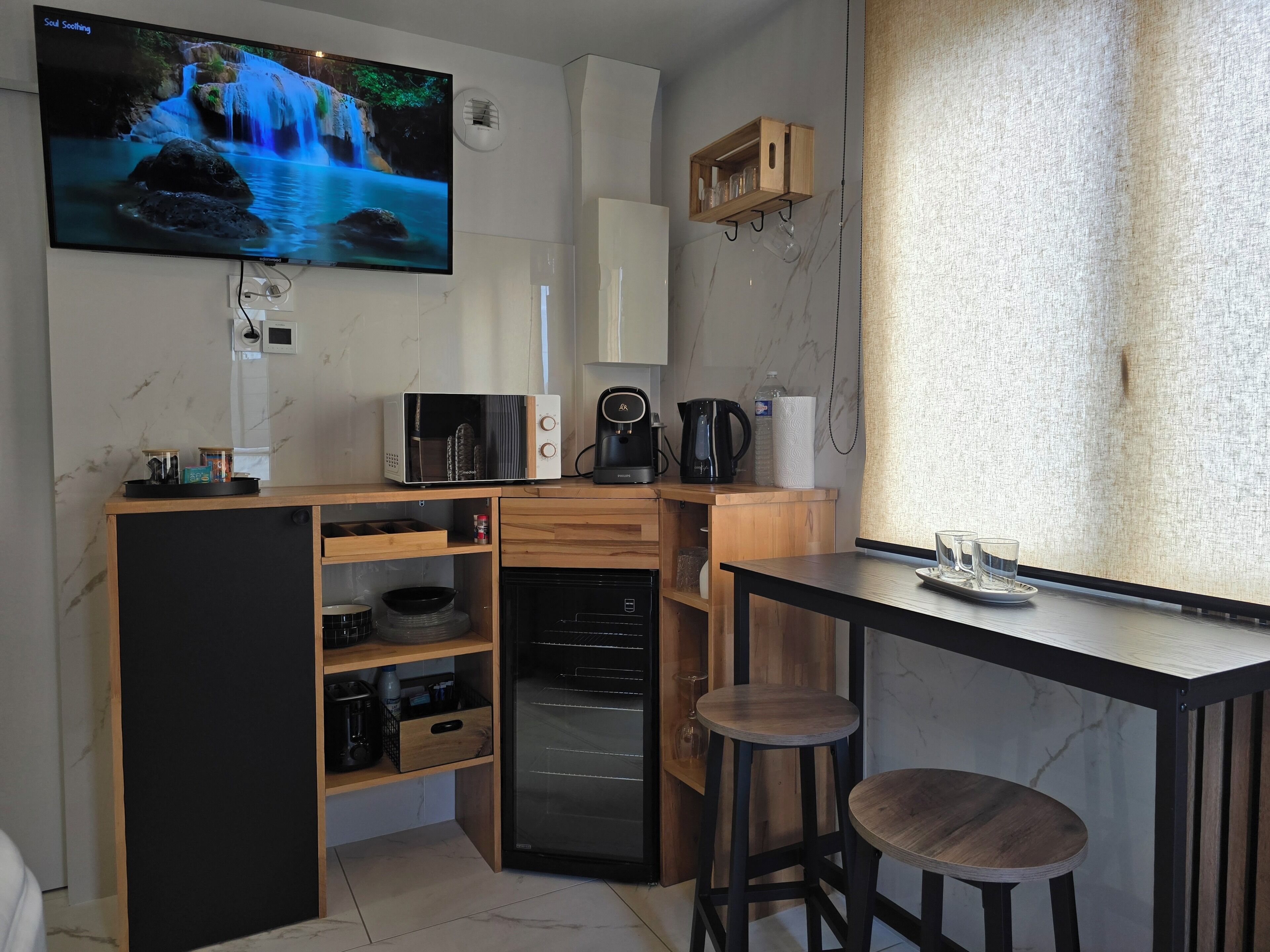 A compact kitchen with a built-in oven, a microwave, a coffee maker, and a TV displaying a waterfall scene.