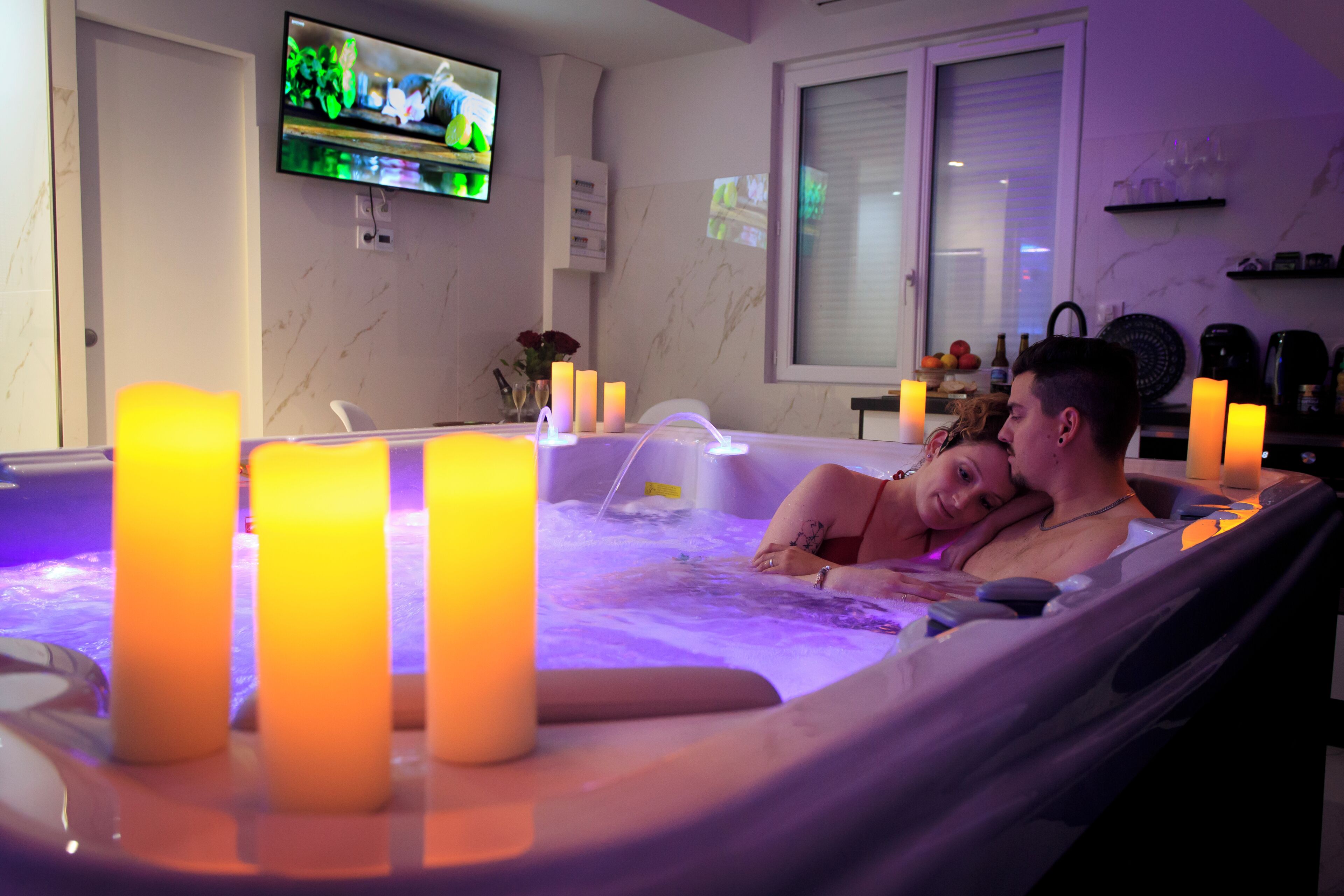 Un couple se détend dans un bain à remous, entouré de bougies, avec une télévision allumée en arrière-plan et une fenêtre munie de stores.