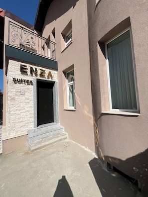Un bâtiment doté d’une façade en pierre, d’une porte d’entrée noire et du nom « ENZA SUITES » au-dessus de la porte.
