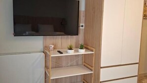 Una habitación con televisión montada en la pared, un armario de madera y una estantería con una planta y un mando a distancia.