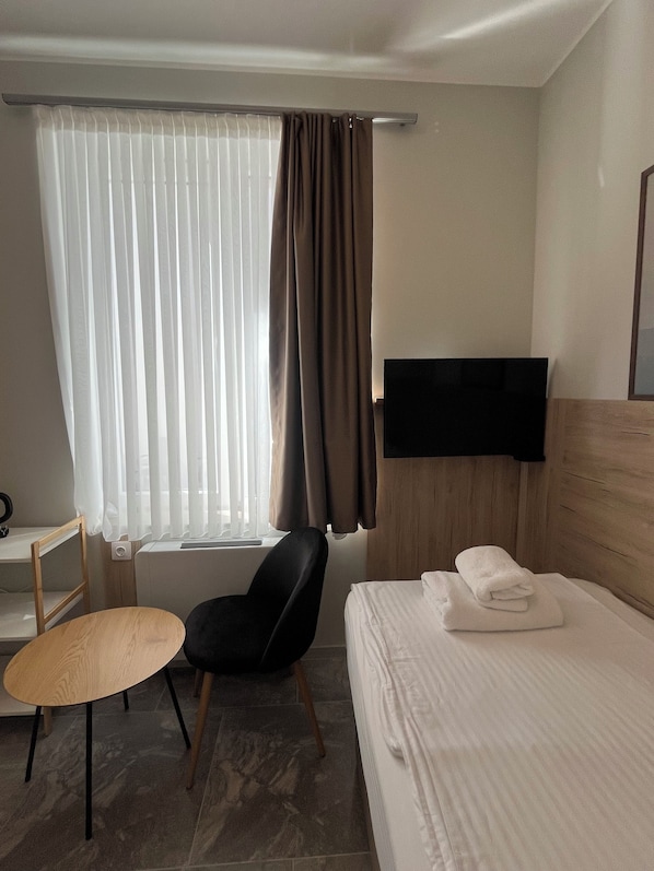 A hotel room with a bed, a chair, a small table, a TV mounted on the wall, and a window with curtains.