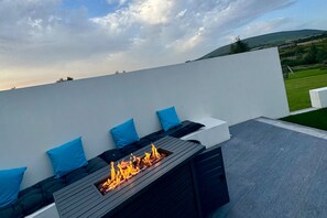 Una terraza en la azotea con una chimenea integrada, asientos con cojines azules y vistas al campo.